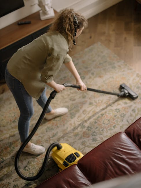 A woman with curly hair, wearing a beige jacket, blue jeans, and white shoes, is performing deep cleaning of a patterned area rug using a yellow vacuum cleaner with a black hose attachment. She is standing on a hardwood floor in a living room, focusing on thoroughly vacuuming the carpet, which shows a detailed floral design with light and dark shades. The room features a wooden console table with a white decorative object on top and a red leather sofa visible in the foreground. The space is well-lit with natural light, emphasizing the cleanliness of the area, and highlights the professional cleaning service provided by Carpet Cleaners Hackney for surface cleaning and maintenance near Mare Street Hackney, HACKNEY.