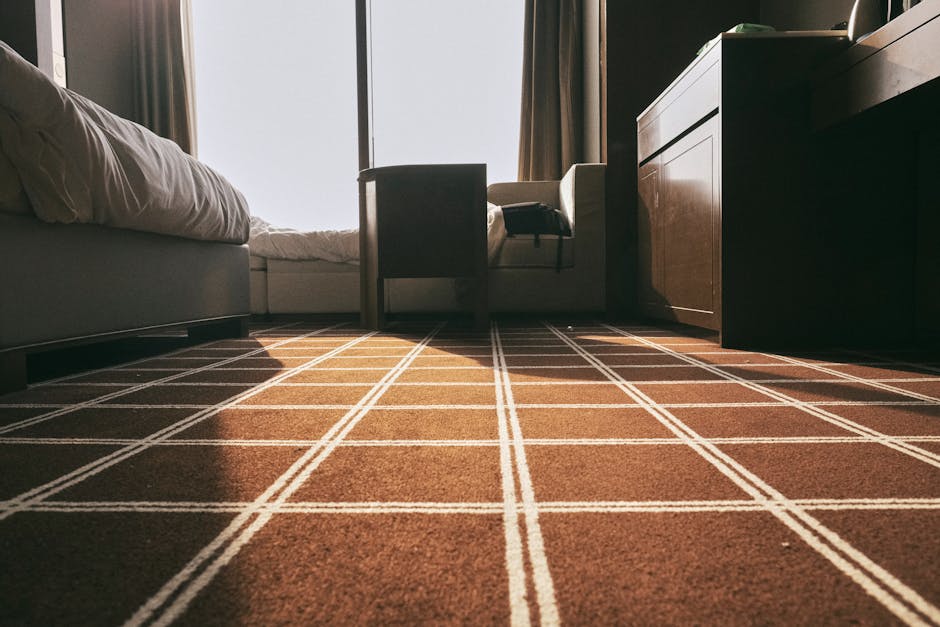 A close-up view of a deep-cleaned living room carpet featuring a brown and white checkered pattern, located in a residential space near Mare Street Hackney. The carpet appears fresh and well-maintained, with a clean, even surface free of dust or debris. In the background, a bed with white linens and a comfortable headboard is partially visible on the left, while a living room sofa and a wooden sideboard are positioned along the right wall. Sunlight filters through a large window with brown curtains, illuminating the room and highlighting the tidy, hygienic appearance of the floor. The image captures a scene indicative of professional surface cleaning, emphasizing the importance of proper deep cleaning and sanitisation, as provided by Carpet Cleaners Hackney, to maintain a pristine home environment.