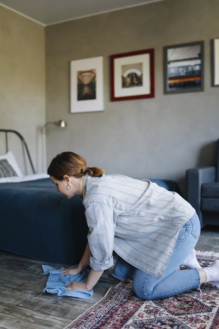A woman with curly hair, wearing a beige jacket, blue jeans, and white shoes, is performing deep cleaning of a patterned area rug using a yellow vacuum cleaner with a black hose attachment. She is standing on a hardwood floor in a living room, focusing on thoroughly vacuuming the carpet, which shows a detailed floral design with light and dark shades. The room features a wooden console table with a white decorative object on top and a red leather sofa visible in the foreground. The space is well-lit with natural light, emphasizing the cleanliness of the area, and highlights the professional cleaning service provided by Carpet Cleaners Hackney for surface cleaning and maintenance near Mare Street Hackney, HACKNEY.
