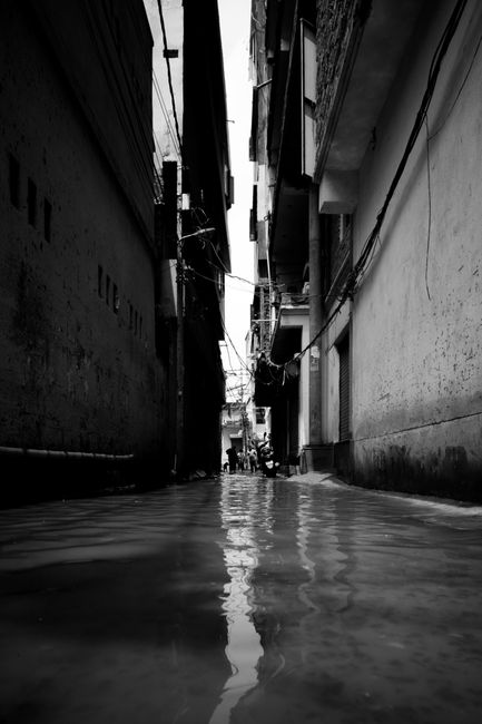 A narrow urban alleyway heavily flooded with water, reaching up to the height of the door sills on both sides. The scene is in black and white, emphasizing the depth of the water which reflects the surrounding buildings. The alley is flanked by multi-story residential or commercial structures with visible windows, balconies, and external wiring. The surfaces of the buildings appear wet and darkened by moisture, with some areas showing signs of water damage. No cleaning tools or personnel are visible in the image. The scene highlights the aftermath of flooding which requires professional emergency water removal and surface cleaning by companies like Carpet Cleaners Hackney, especially in areas affected by sudden water ingress such as Clapton Common, HACKNEY.