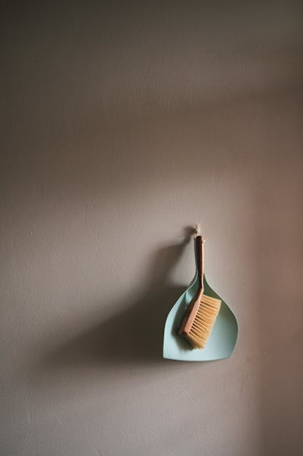 A light blue wall hook mounted on a neutral-colored wall in a residential space holds a brown-handled cleaning brush with natural bristles. The brush rests inside a matching light blue, semi-circular wall-mounted holder, which appears to be made of plastic or ceramic. The wall's surface is smooth and clean, with soft, natural lighting illuminating the area, emphasizing the tidy and minimal aesthetic. The arrangement suggests an organized space dedicated to home cleaning supplies, aligning with the themes of surface cleaning, domestic hygiene, and maintenance, as offered by Carpet Cleaners Hackney for end of tenancy cleaning checklists and quotes in Hackney Central.