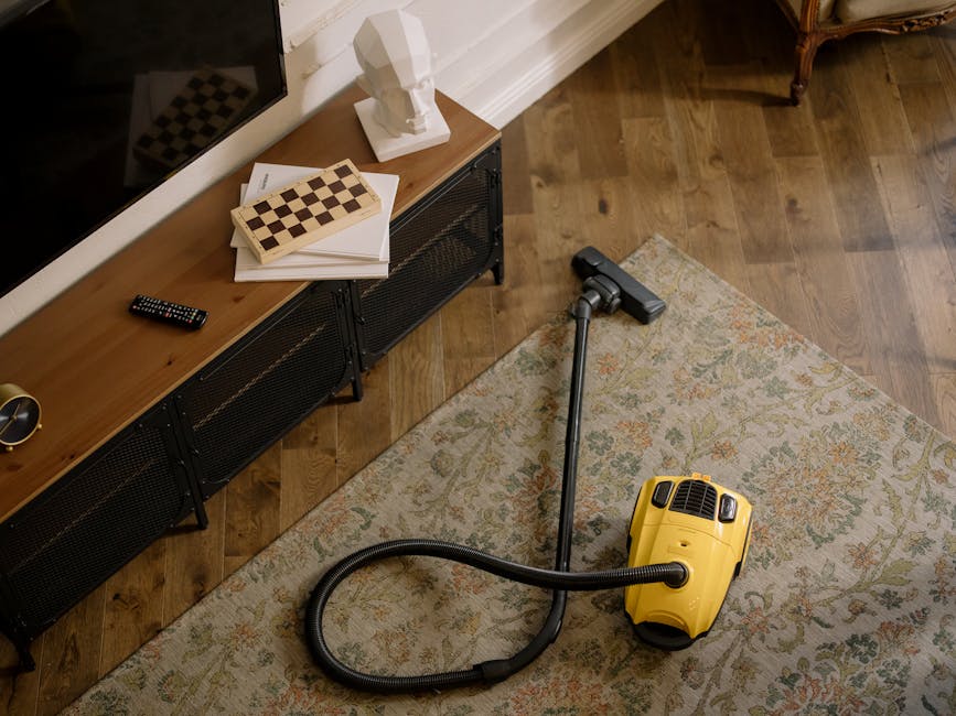 A person kneeling on a patterned carpet in a room, operating a yellow portable carpet cleaning machine with a black hose. The individual is wearing a khaki jacket and blue jeans, and is in the process