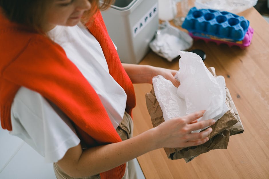 A person wearing a white shirt with a red scarf around their shoulders is holding a crumpled piece of white tissue paper and a brown paper bag, standing next to a wooden table. On the table, there is 