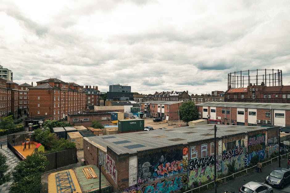 A wide view of an urban area in Hackney, showing a cluster of low-rise brick buildings, some with graffiti-covered walls and rooftop structures, under a cloudy sky. The scene includes a mix of residential and industrial-style surfaces, with paved roads and a few parked cars. Some small trees and greenery are visible among the buildings, and the overall atmosphere suggests a well-maintained but naturally weathered environment. The image captures the architectural layout and street-level details of the area, consistent with the aesthetic of Hackney Wick. Carpet Cleaners Hackney's branding or cleaning tools are not visible in this outdoor scene, but the description aligns with the company's expertise in surface cleaning and maintenance relevant to urban settings.