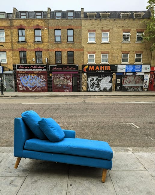 A bright blue modern chaise lounge with matching cushions, positioned on the sidewalk in front of a row of shopfronts adorned with graffiti and posters in Hackney Wick. The furniture appears clean and well-maintained, highlighting the surface cleaning and maintenance services offered by Carpet Cleaners Hackney. The background features a multi-story brick building with windows and signage for various businesses. The scene is illuminated by natural daylight, emphasizing the contrast between the clean upholstery and the urban environment. This image illustrates the importance of professional deep cleaning and sanitisation for domestic and commercial furnishings, as part of thorough hygiene and upkeep routines.