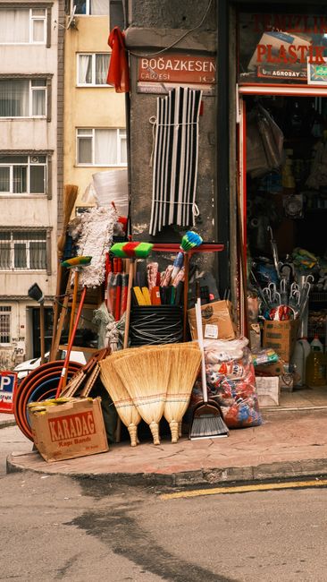 A person wearing yellow rubber boots and casual clothing is engaged in surface cleaning outside a commercial shop in Dalston, Hackney, during the daytime. The individual is using a floor scrubber or p