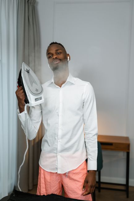 A man with dark skin and short hair is standing indoors, holding a steamer iron emitting steam in his right hand. He is dressed in a white shirt and pink patterned trousers, with a neutral expression on his face. The room features a white wall with gray curtains on the left side, and a wooden desk with black legs is visible in the background. The lighting is soft and natural, creating a clean and tidy atmosphere suitable for domestic surface cleaning or deep sanitisation efforts, reflecting the focus of the page on steam cleaning tips for Broadway Market flats and landlords, as provided by Carpet Cleaners Hackney.