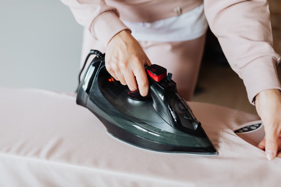 Close-up of a person using a black and red steam iron on a light beige fabric surface, likely upholstery or curtains, in a well-lit room. The person's hand is pressing down on the iron's handle while the iron is moved smoothly across the fabric, which appears clean and free of wrinkles. The background is slightly blurred, emphasizing the ironing process. This image illustrates domestic surface cleaning and maintenance, relevant to deep cleaning tips for Broadway Market flats and landlords as promoted by Carpet Cleaners Hackney.